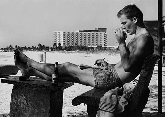Hunter S. Thompson sitting down on a chair in the beach smoking a cigarette and writing