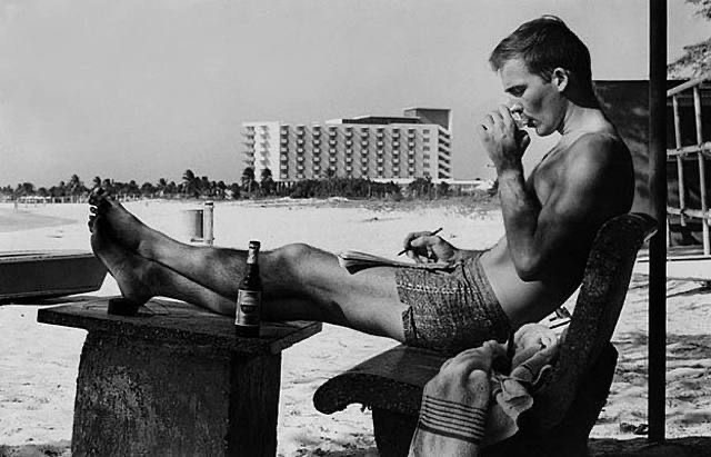Hunter S. Thompson sitting down on a chair in the beach smoking a cigarette and writing